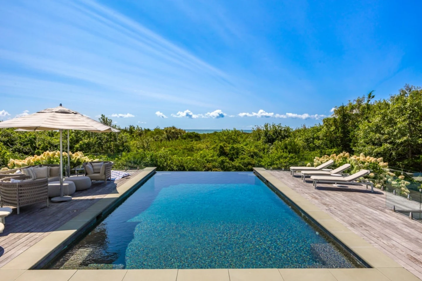 Infinity edge pool overlooking dense greenery, deck seating, and sunny sky