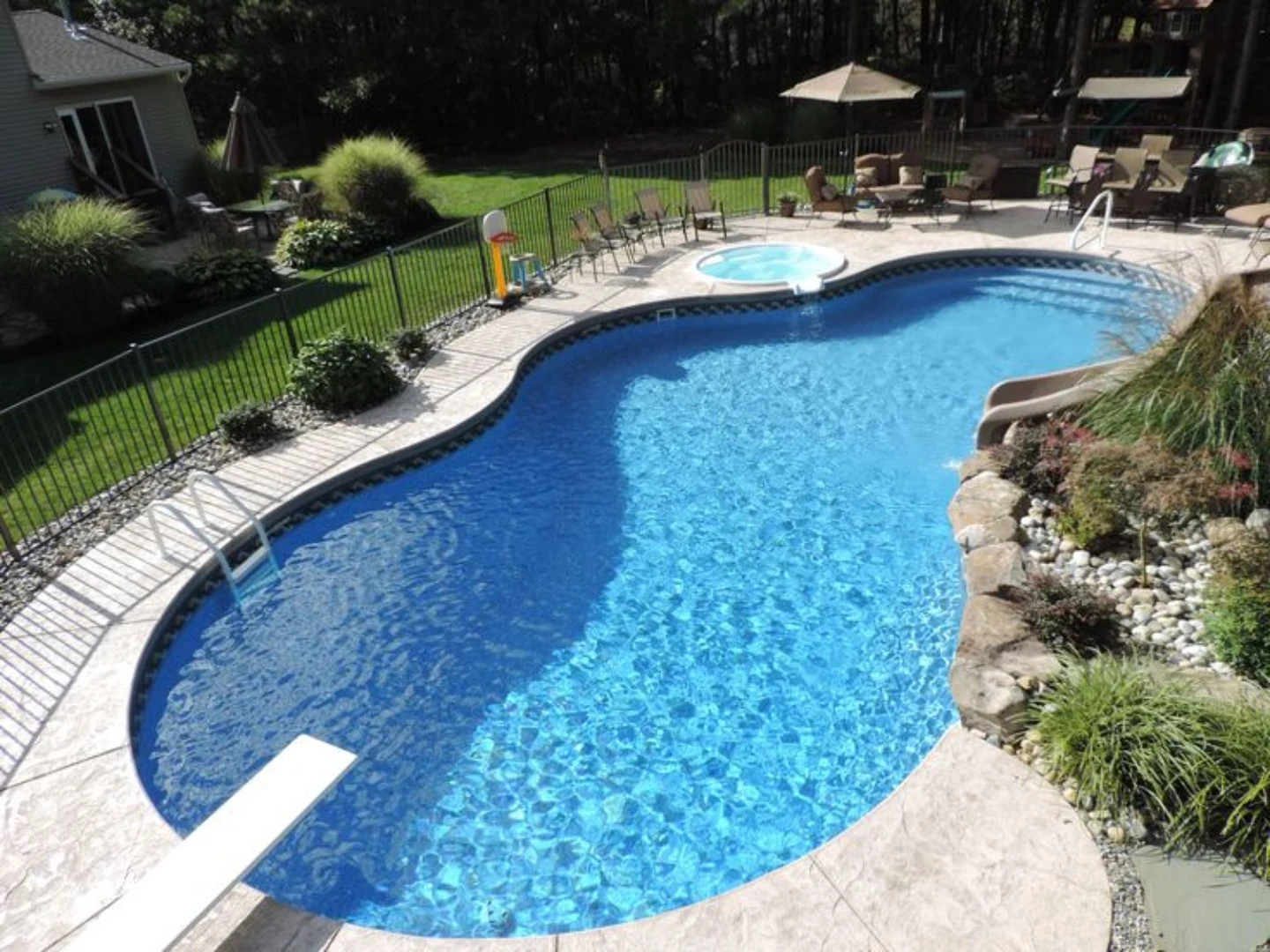 Curved backyard pool with spa, diving board, slide, and surrounding fence