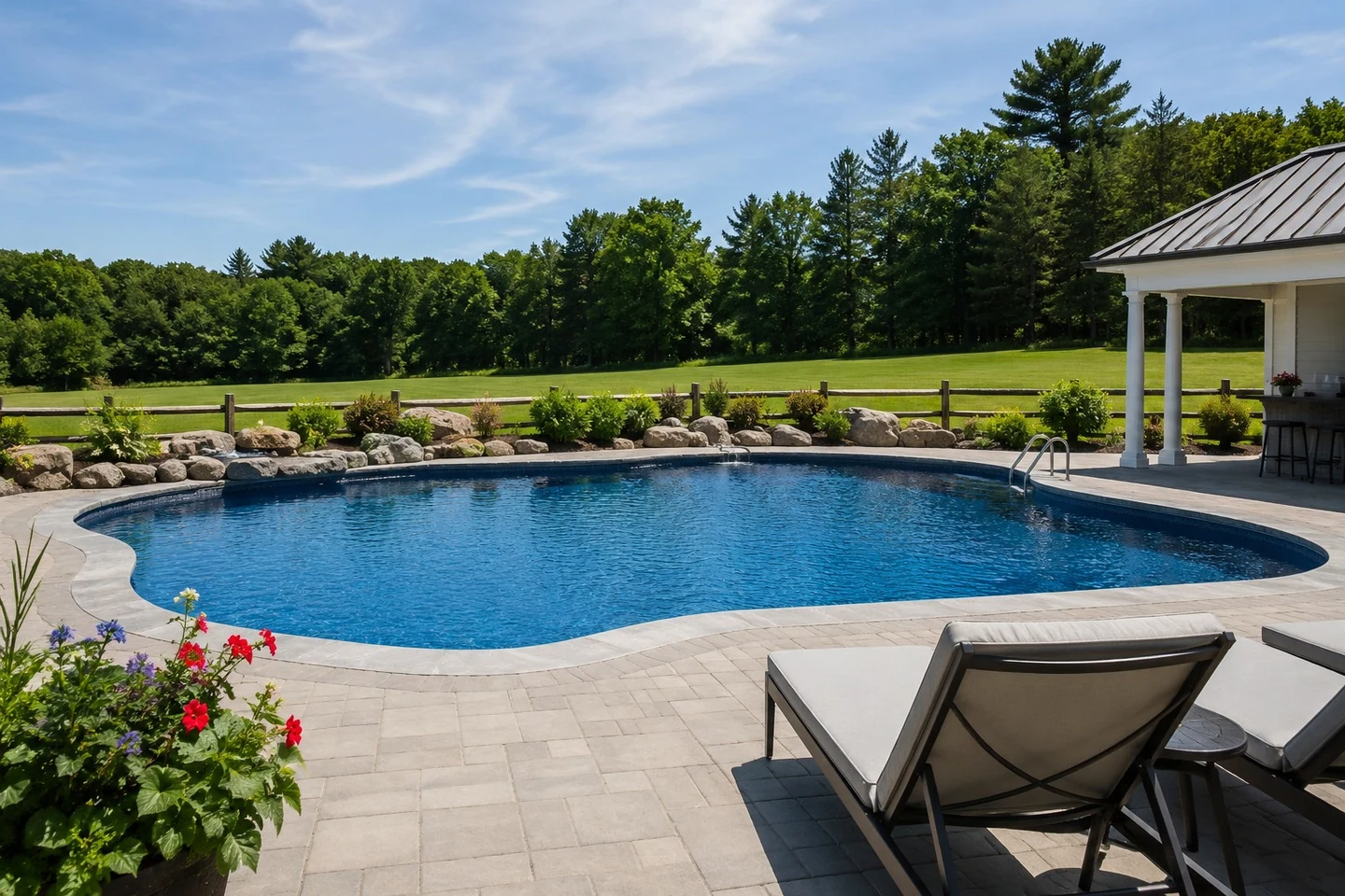 Freeform backyard pool with paver patio, loungers, and rock border facing field