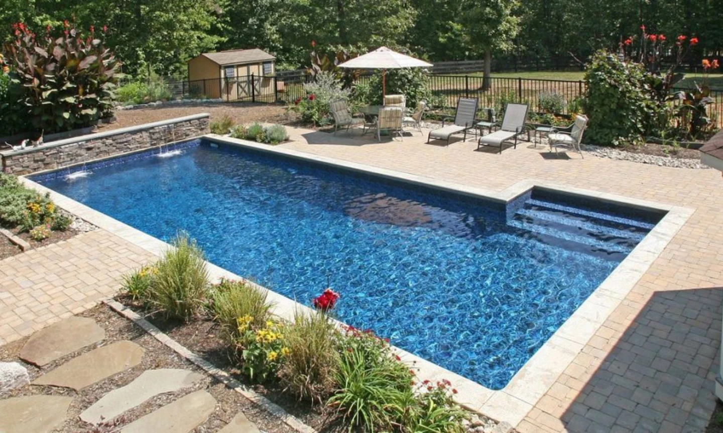 Rectangular blue pool with stone coping, steps, patio furniture, and planting