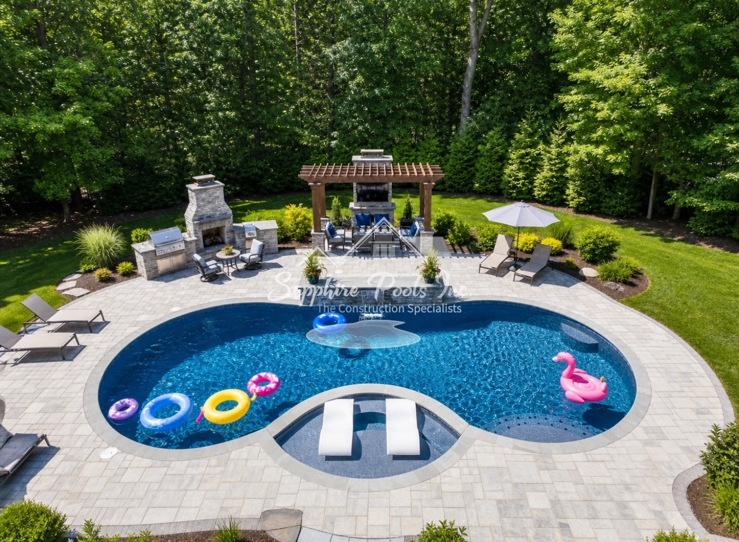 Aerial daytime view of a vinyl-liner pool, patio, pergola, outdoor kitchen, landscaping, and colorful pool floats